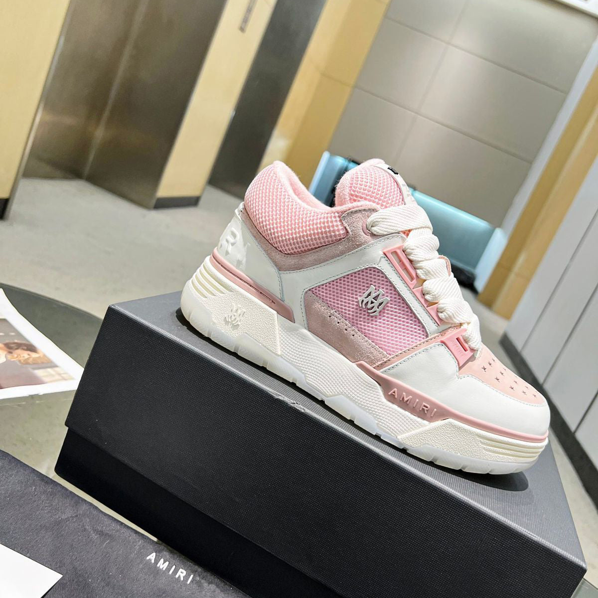 Pink and white sneaker on a black box with 'AMIRI' branding in an indoor setting.